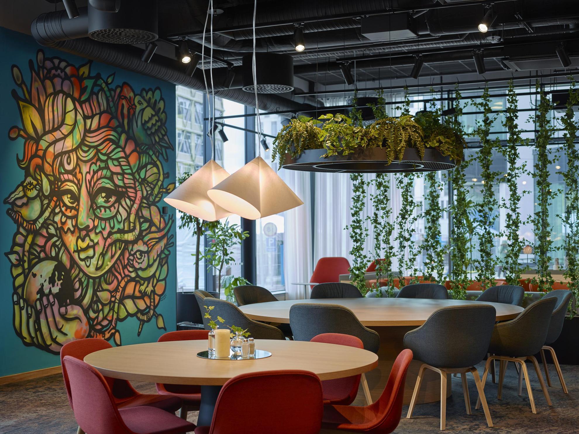 Een kamer met ronde houten tafels met rode en zwarte stoelen. Aan het plafond hangen planten en een blauwe muur is beschilderd met kunstwerken.
