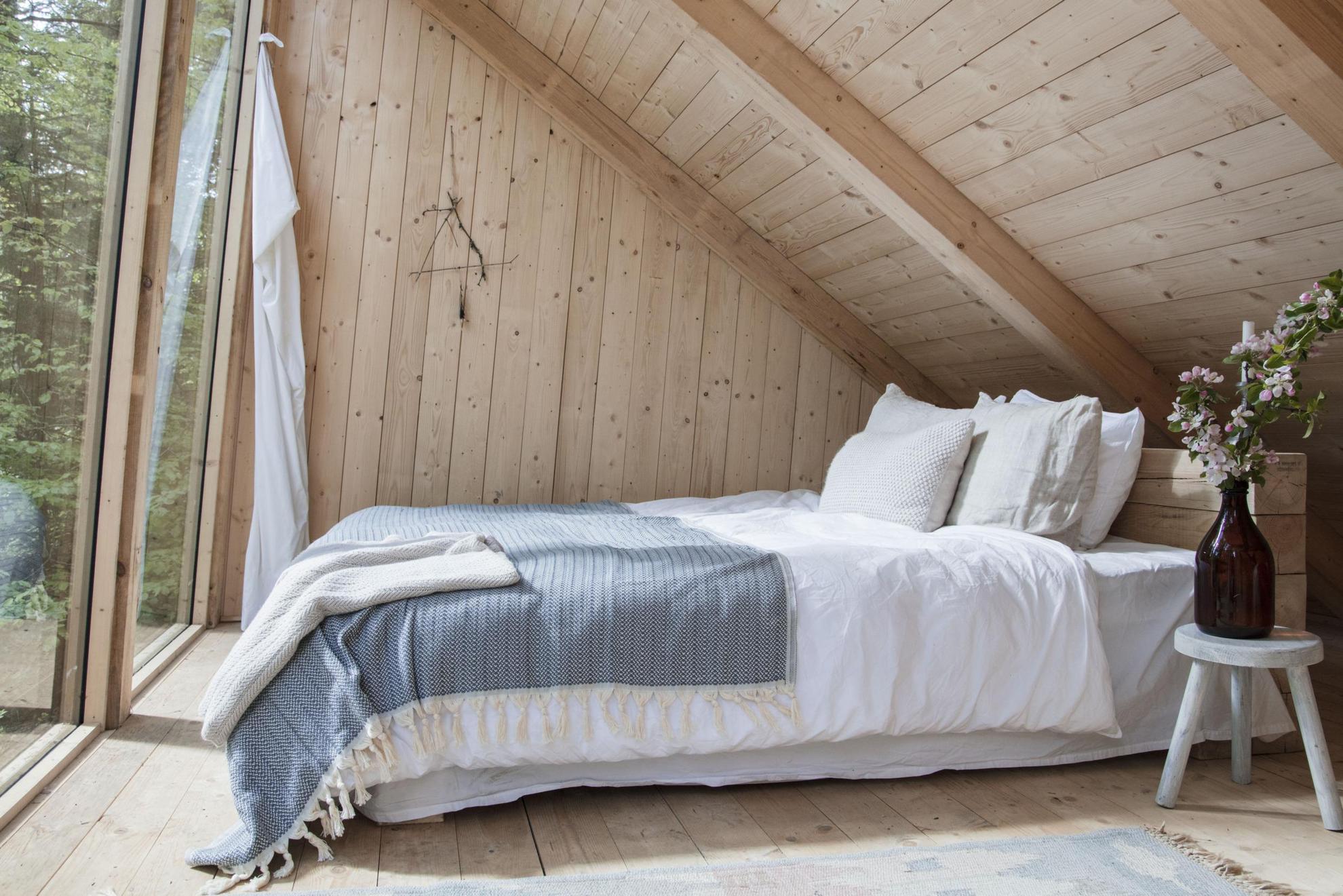 Een kleine hut met houten wanden, een schuin plafond en kamerhoge ramen. Een knus tweepersoonsbed met wit beddengoed en een deken in het midden van de kamer.