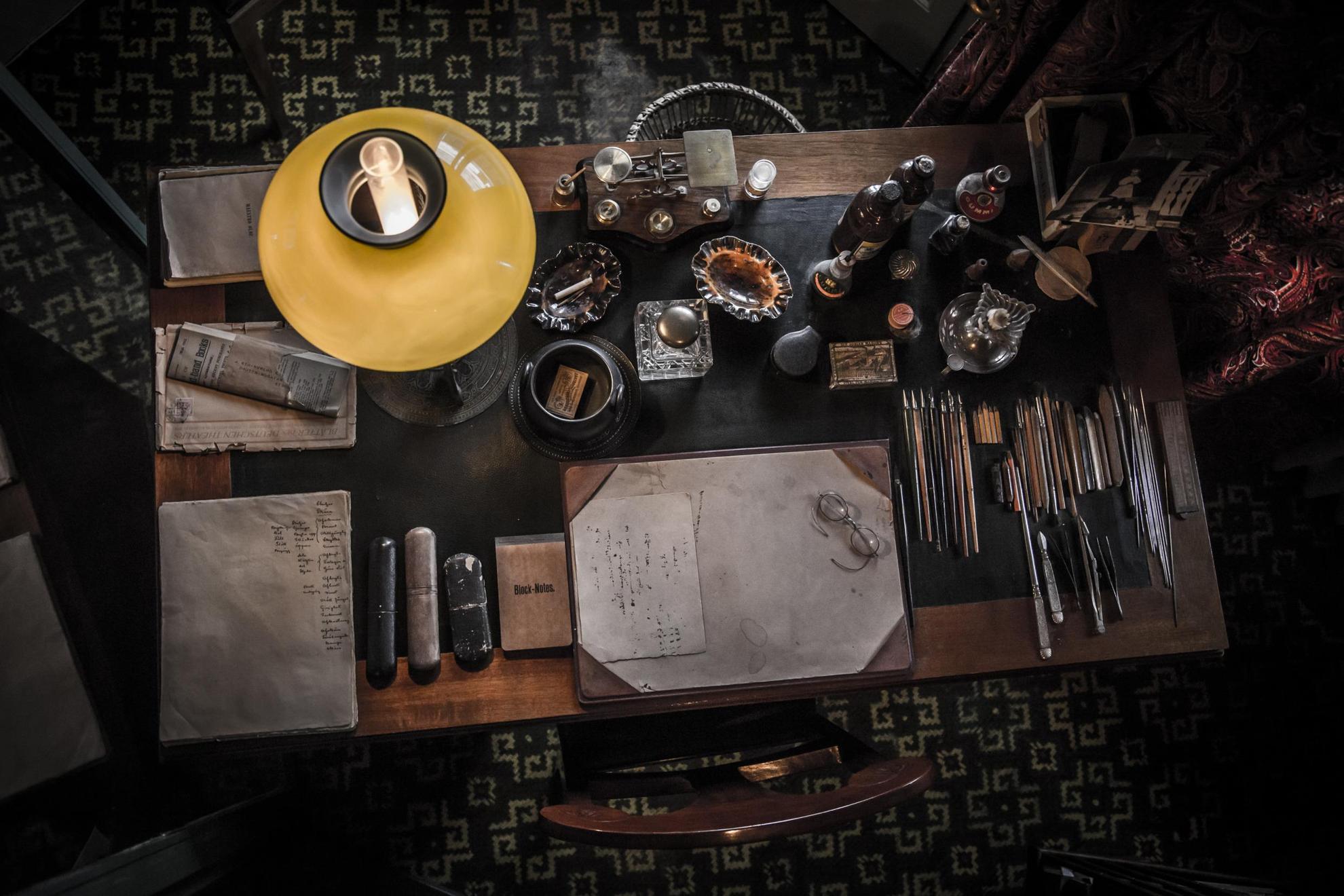 Een oud houten bureau, met potloden in een rij, papier en notities, een bril en een oude lamp.