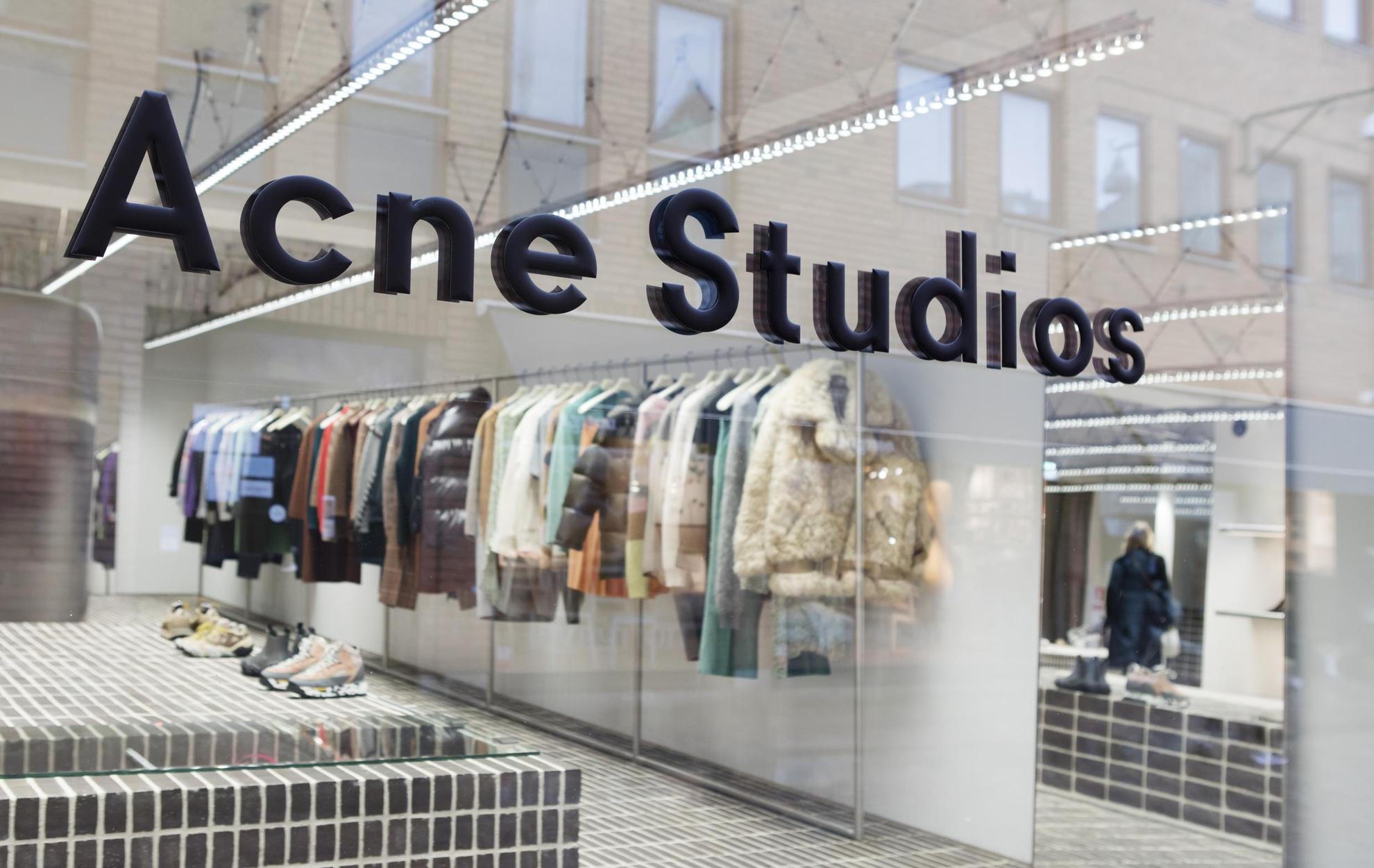 Een etalage met een bord waarop Acne Studios staat. Binnen zie je een rek met verschillende kleding en wat schoenen.