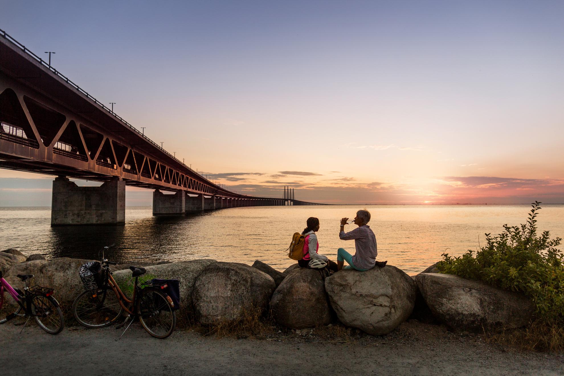 Twee vrouwen zitten bij zonsondergang bij de Öresundbrug in Malmö en drinken water uit een fles, twee fietsen geparkeerd op de grindweg.