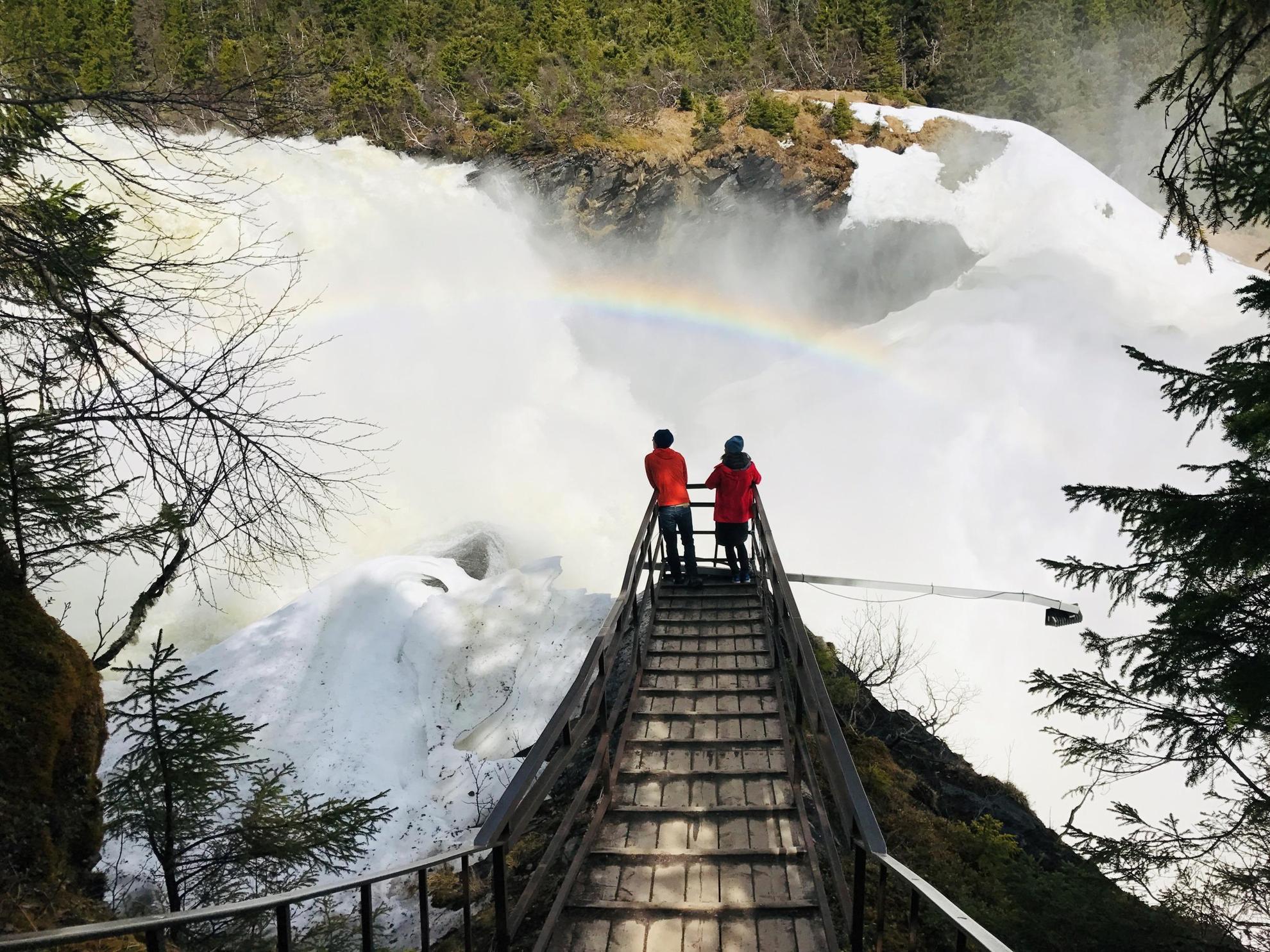 Twee mensen kijken uit op een enorme waterval. Het water van de waterval creëert een regenboog.