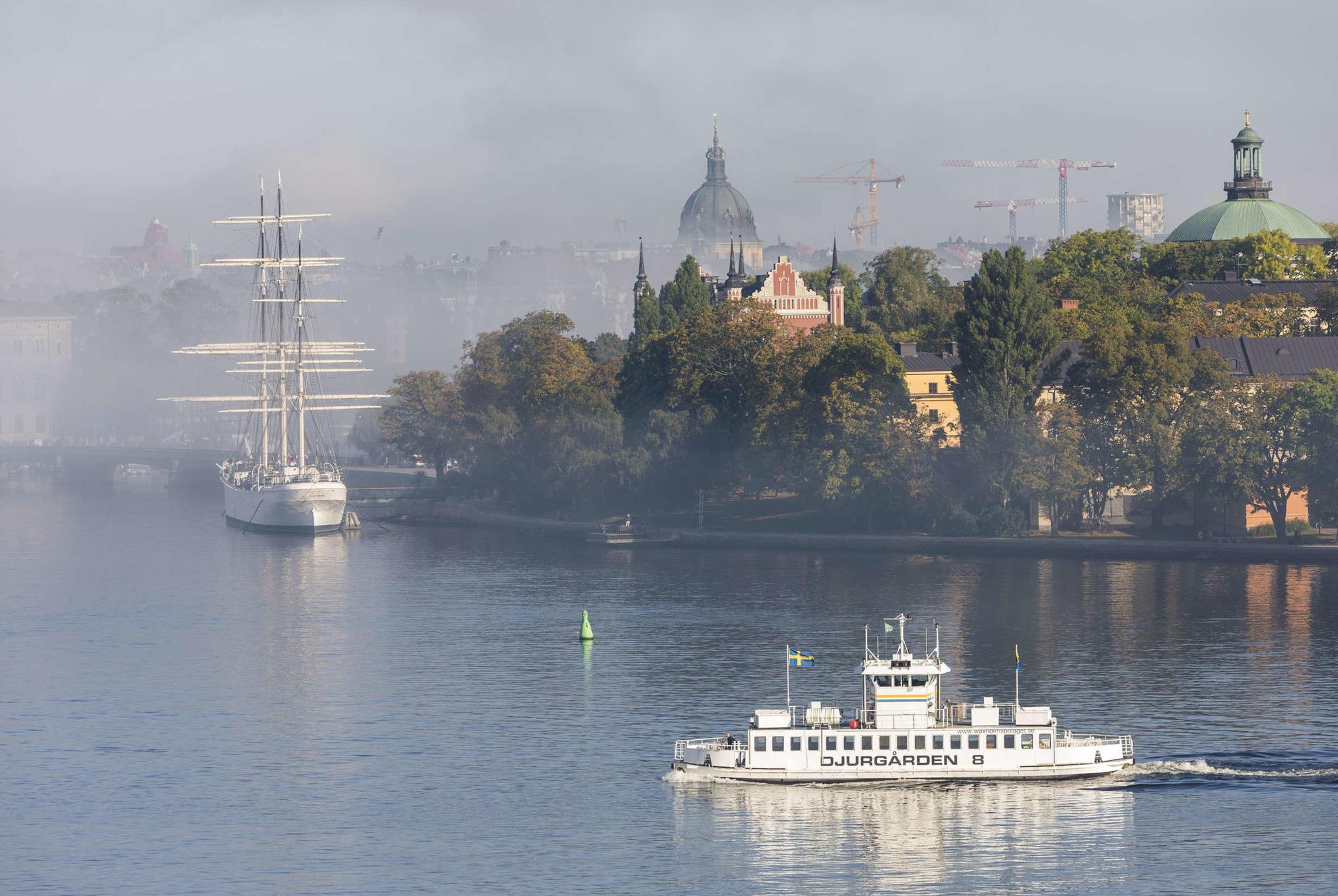 Een openbaarvervoerboot die op een mistige zomerdag het eiland Kastellholmen passeert, met groen en grote gebouwen.