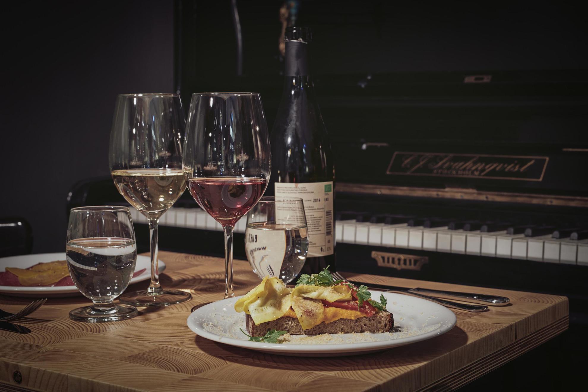 Een gedekte tafel met een fles wijn, twee borden met eten en twee glazen wijn. De tafel staat naast een piano.
