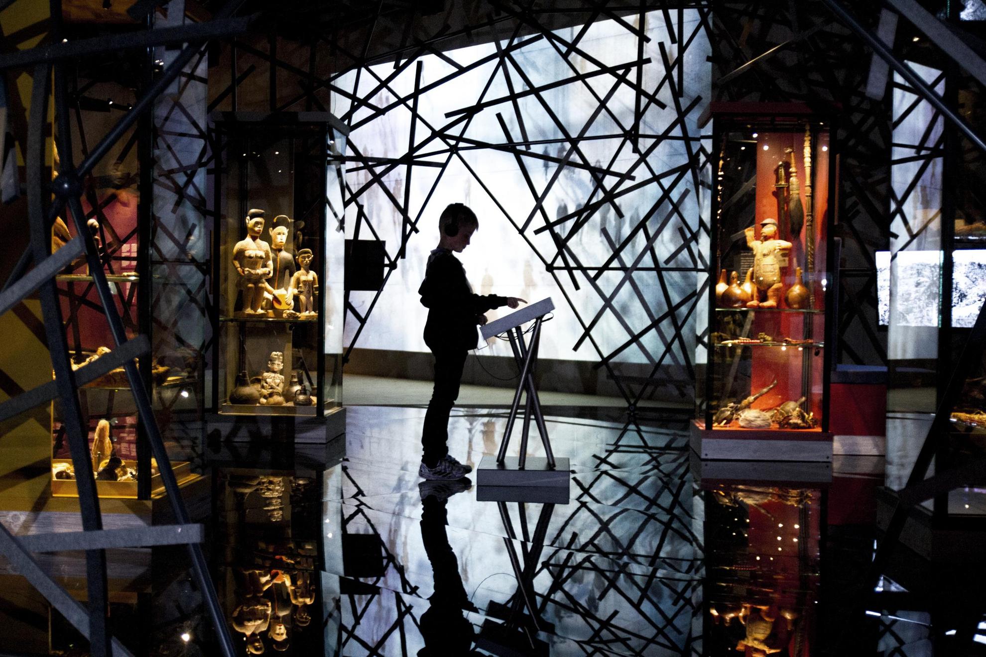 Een kind staat voor een display in het midden van een tentoonstellingsruimte. De ruimte is donker maar verlicht door een groot scherm achterin, omgeven door kruisende stokken, wat een dramatisch effect geeft met het silhouet van de bezoeker in het midden.