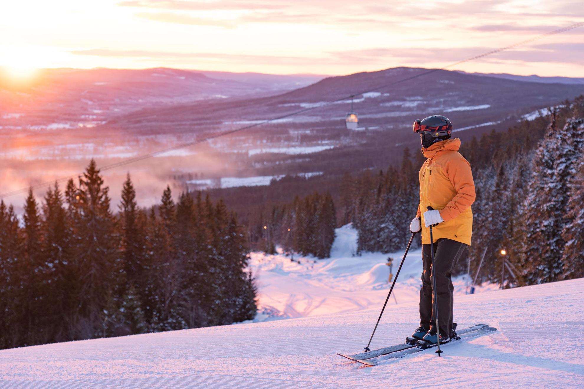 Een persoon met skiuitrusting staat op een skihelling en kijkt naar de zonsondergang.