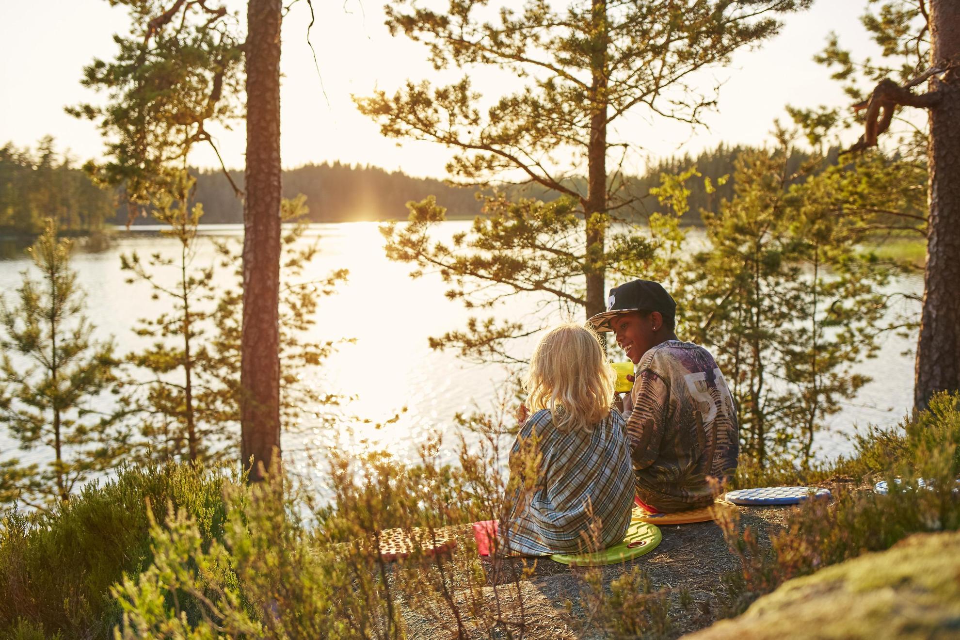Twee jonge personen zitten op een kleurrijk kussen op de rotsen over het water uit te kijken in een bosrijke omgeving.