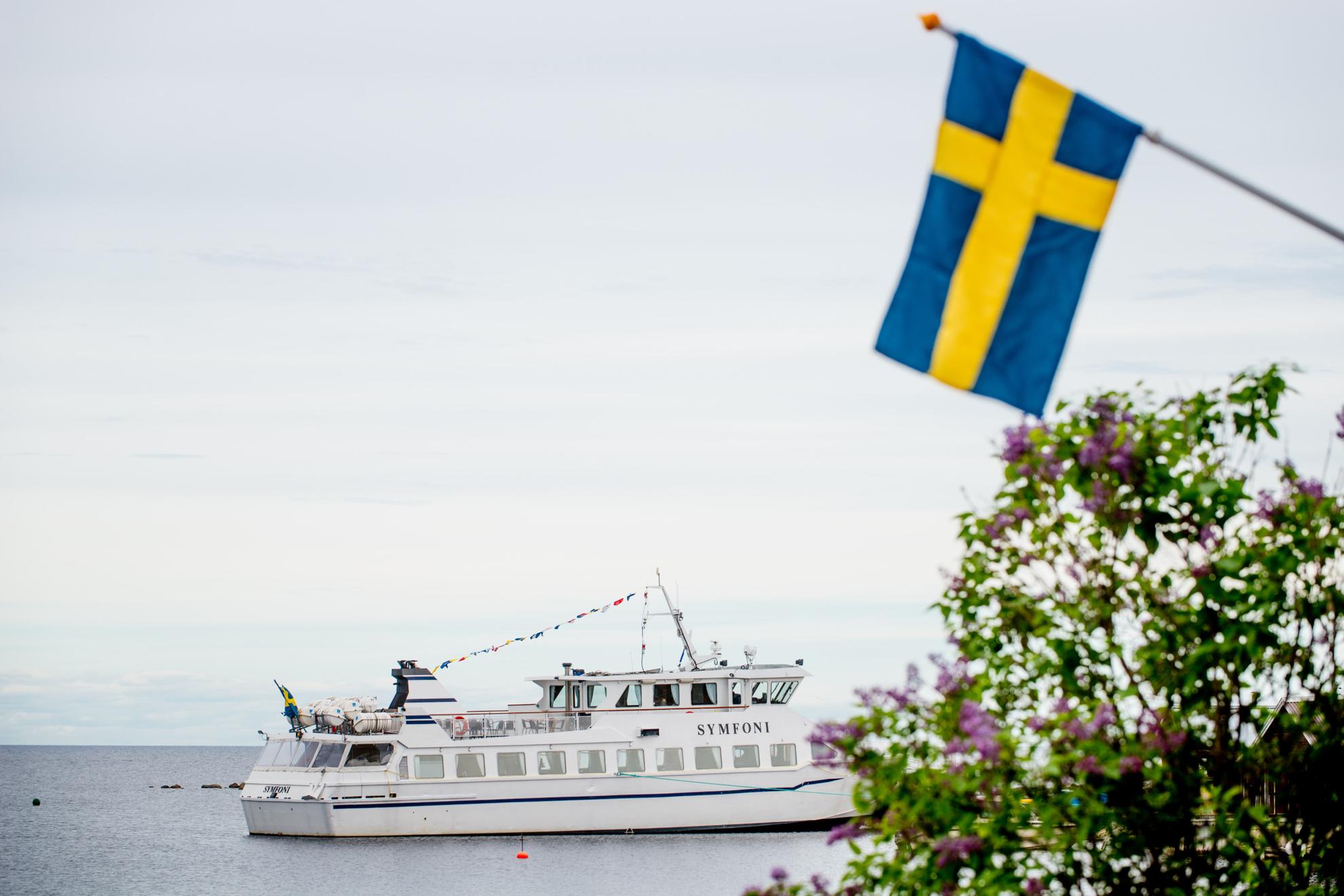Een boot genaamd Symfoni drijft in het water, met een Zweedse vlag en paarse bloemen op de voorgrond.