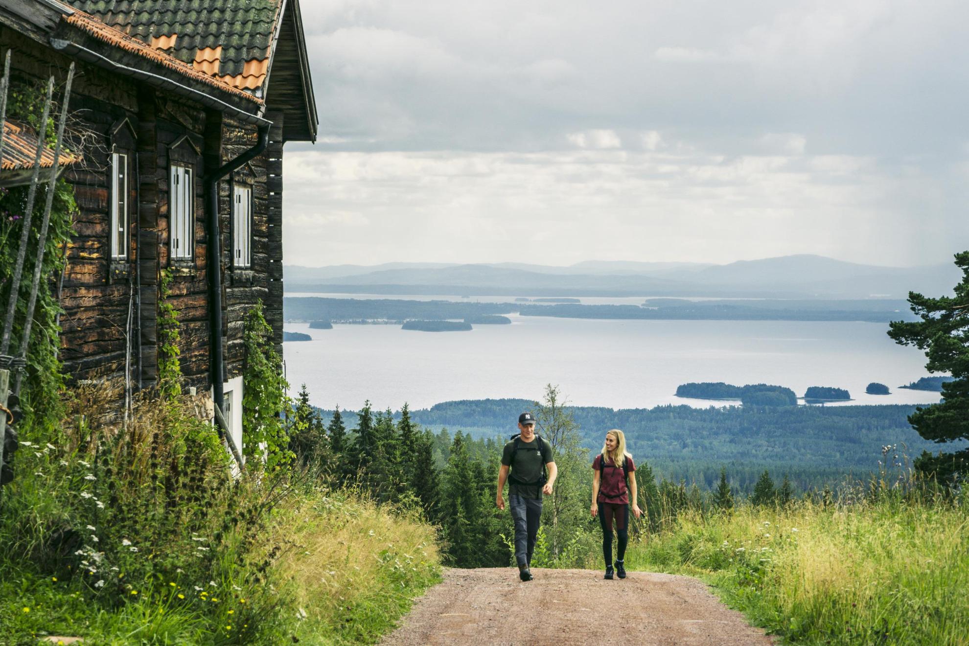Een stel wandelt op een heuvel naast een houten huis met op de achtergrond uitzicht op een meer.