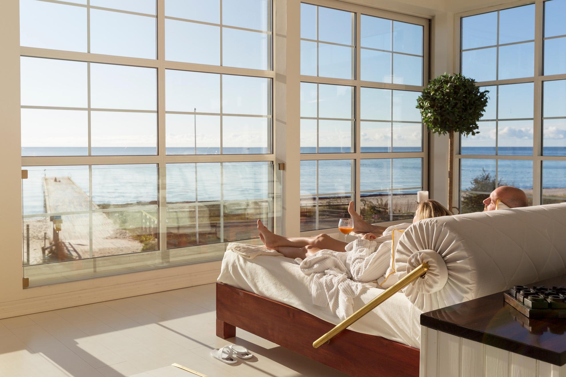 Een stel ligt in een groot bed naast grote ramen met uitzicht op het strand en de zee