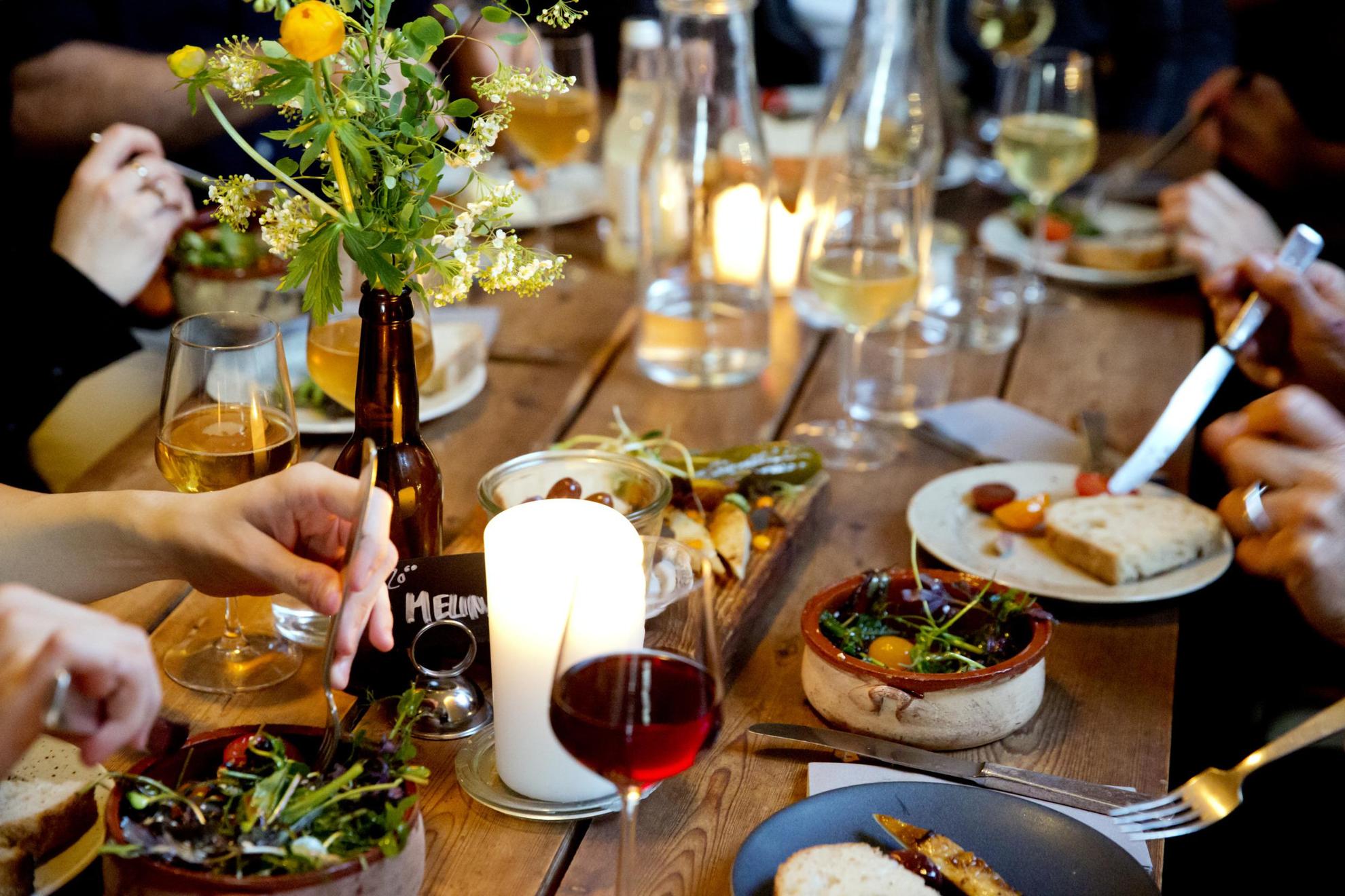 Een houten tafel waarop kleine vegetarische gerechten staan. Iemand schept salade van een bord op. Rondom de tafel staan wijnglazen en kleine witte borden.
