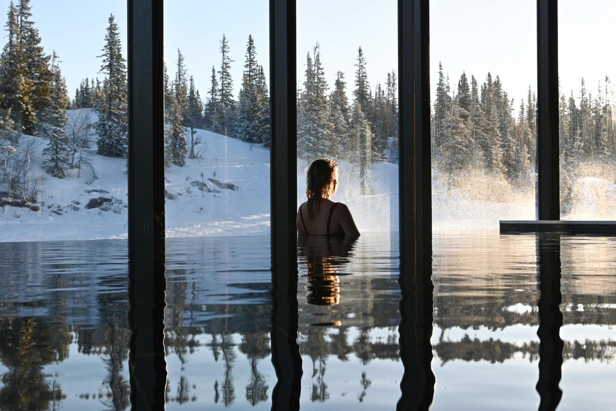 Een persoon in een overdekt overloopzwembad dat door de grote ramen uitkijkt op het met sneeuw bedekte bos.