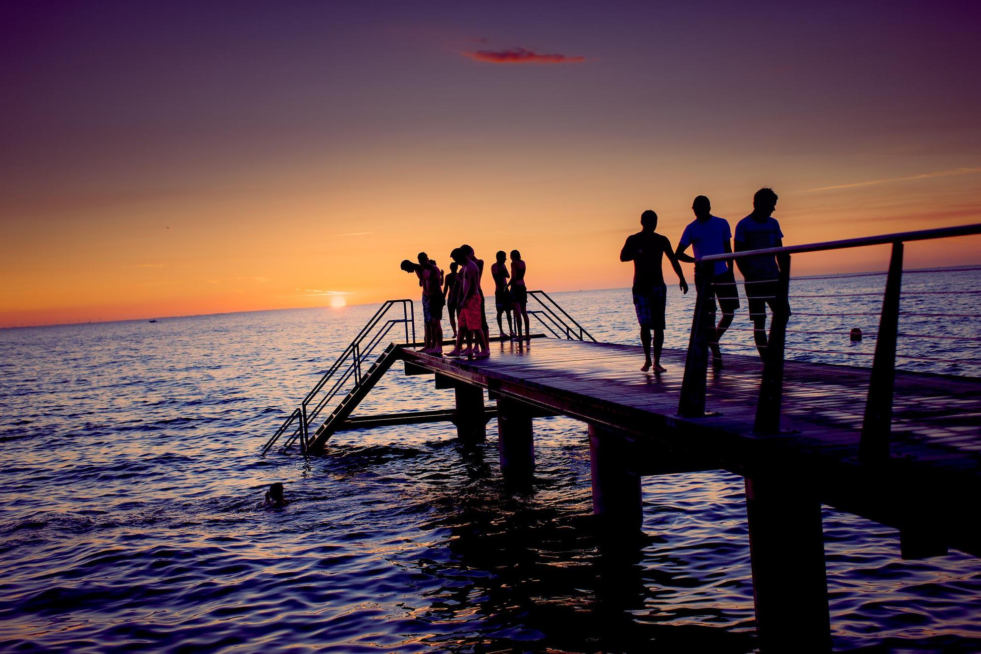 Een groep mensen staat tijdens zonsondergang op een pier in Zweden. Sommigen kijken naar een persoon die aan het zwemmen is.