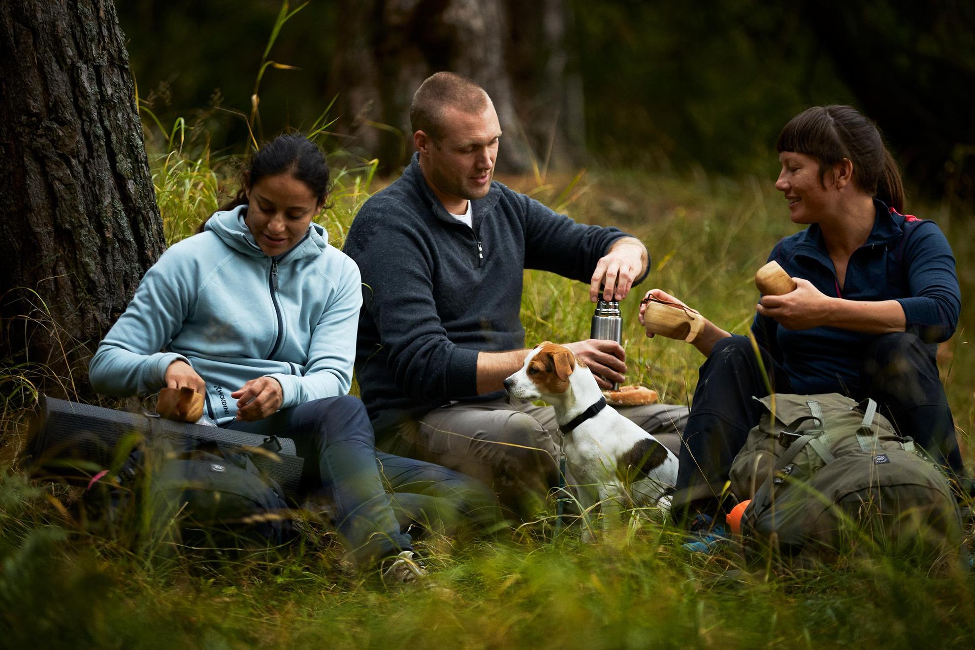 Drie personen zitten op de grond in een bos en drinken koffie uit een thermos.