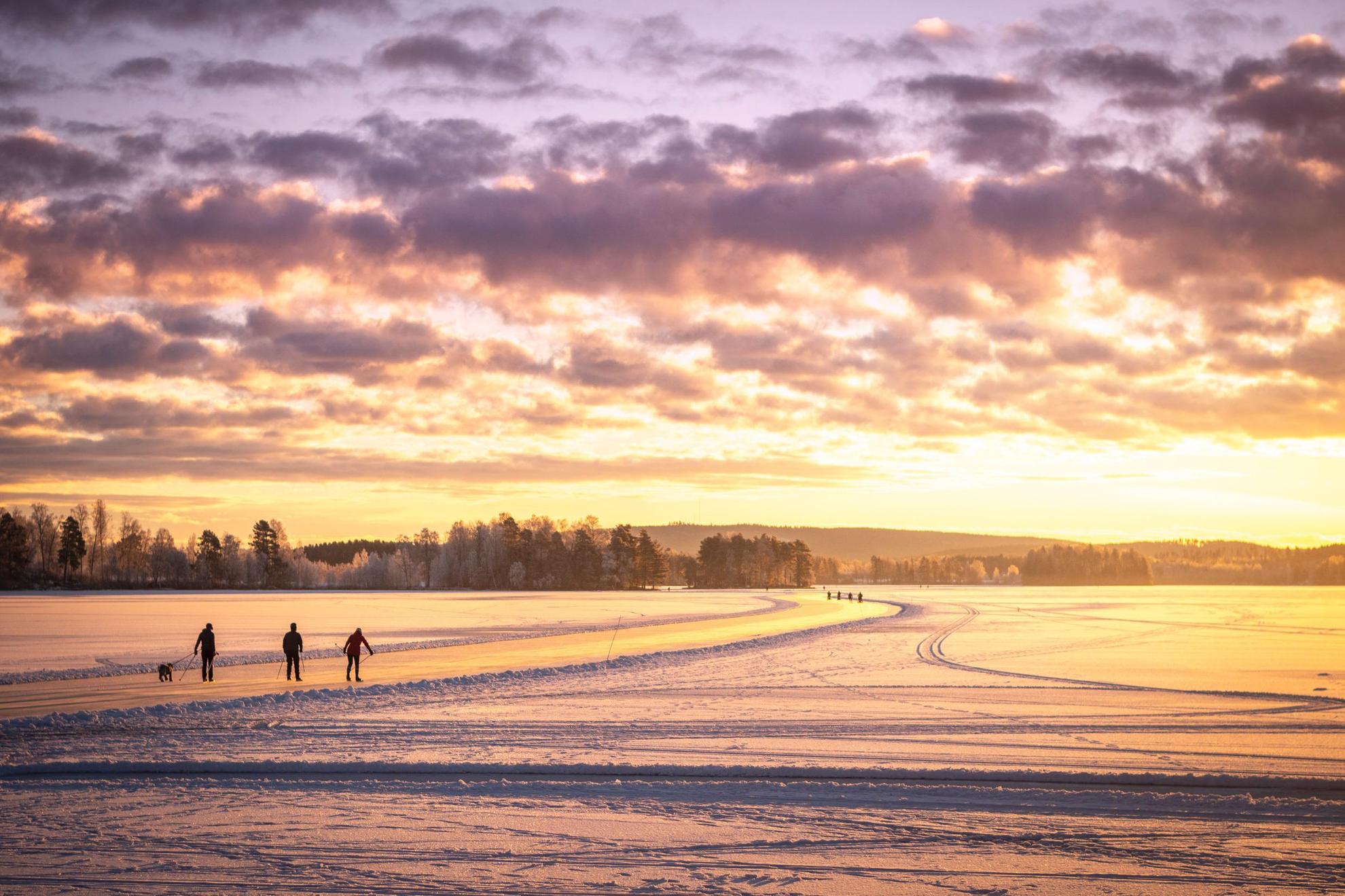 Silhouet van drie mensen die schaatsen op een bevroren meer. De zon schijnt oranje.
