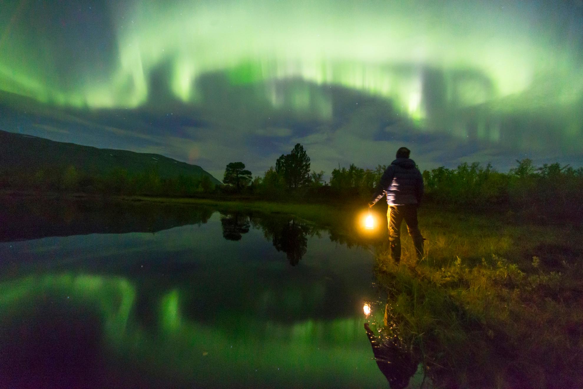 Een persoon die een lamp vasthoudt, kijkt naar het groene noorderlicht aan de hemel. Het noorderlicht reflecteert in het meer naast de persoon.