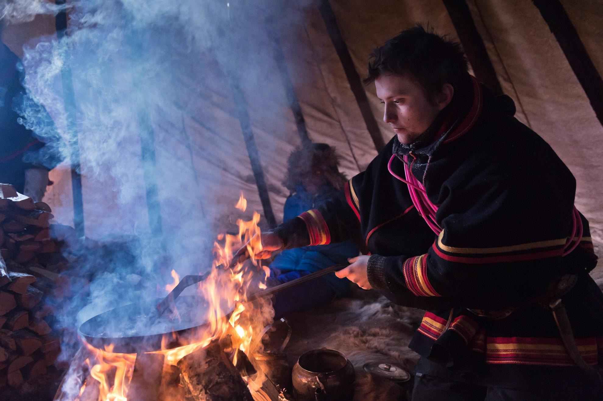 Een persoon in Sami klederdracht kookt boven een vuur in het donker.