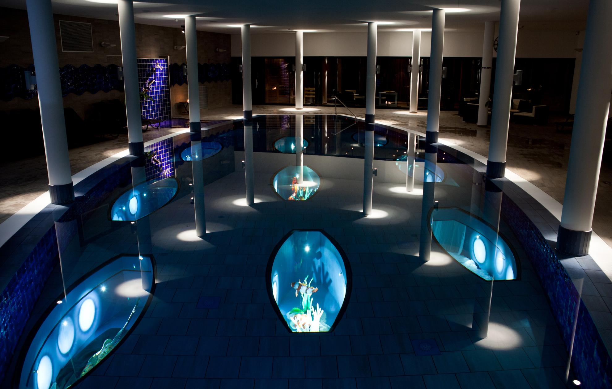 Een binnenzwembad waar het licht gedimd is. In het zwembad zijn verlichte kunsttentoonstellingen met glazen vissen.