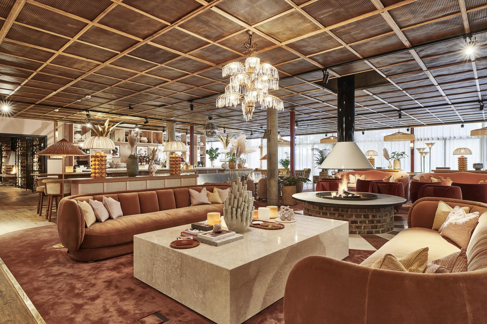 Een loungehoek gedecoreerd met banken en kussens in beige, bruin en lichtroze. In het midden van de kamer is een open haard.