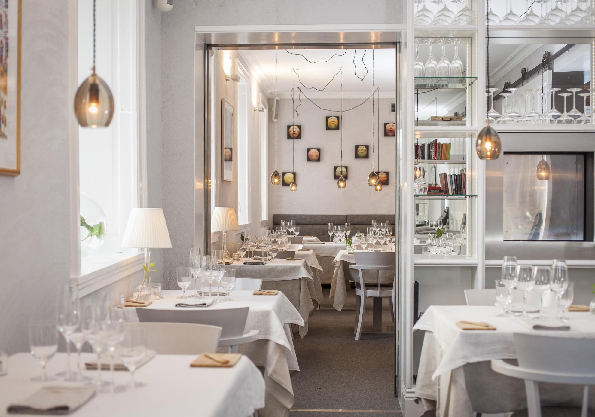 In een restaurant met witte muren zijn de tafels gedekt met witte tafelkleden, linnen servetten en wijnglazen. Aan het plafond hangen lampen.